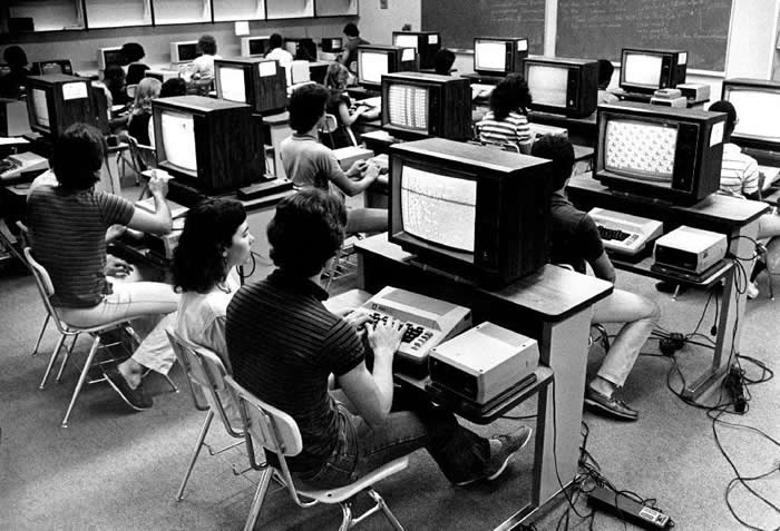 A computer lab in 1985 with at least twelve Atari 800s, Atari's high-end 8-bit computer first released in 1979. Each station includes an 810 disk drive and a large TV monitor. Along the back wall sits a line of Apple IIs, completing the classic setup of the era.