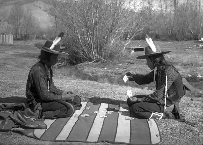 Two Native American men on the Flathead Indian Reservation in western Montana sit on a striped blanket and play cards in the early 1900s.