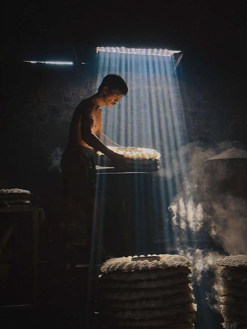 A worker stands indoors beside steaming trays, illuminated by powerful rays of sunlight streaming through a roof opening. The dark surroundings contrast with the bright beams and drifting steam, creating a dramatic, atmospheric scene rich in texture and emotion.