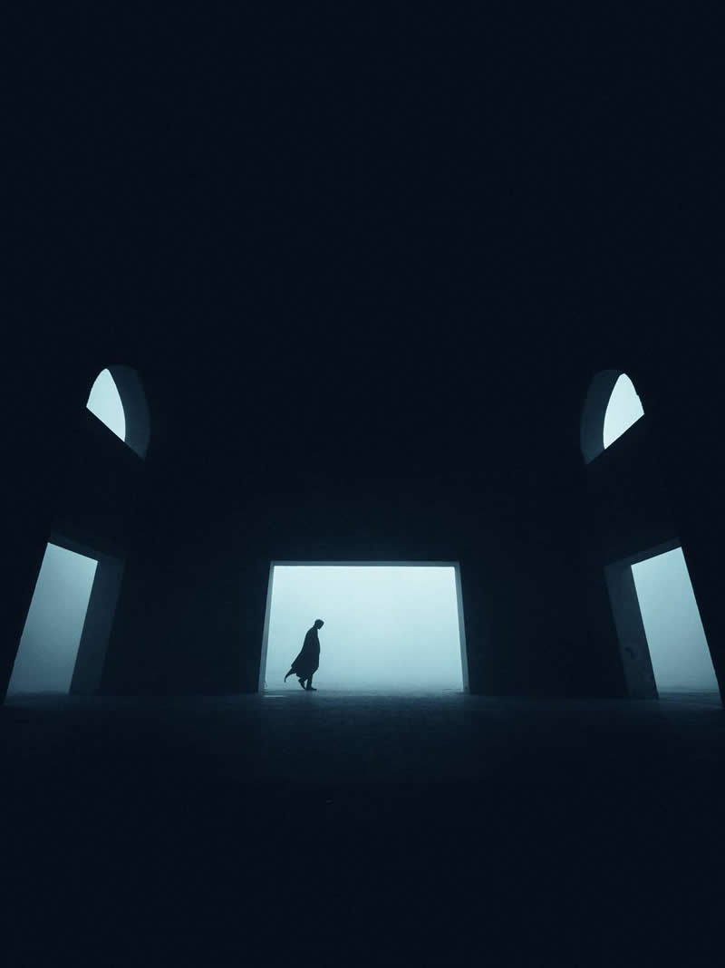 A lone silhouetted figure walks through a dark architectural space toward a bright, fog-filled opening. Symmetrical archways and strong contrast between shadow and light create a minimalist, moody scene with a sense of mystery, scale, and solitude.