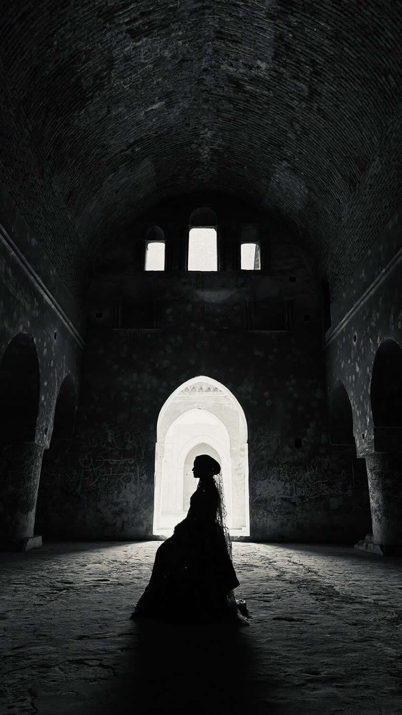 A silhouetted figure in a long dress sits inside a large historic hall, framed by bright arched doorways. Dramatic contrast between the dark interior and glowing background emphasizes the architecture, symmetry, and the subject&rsquo;s graceful profile, creating a moody, elegant atmosphere.