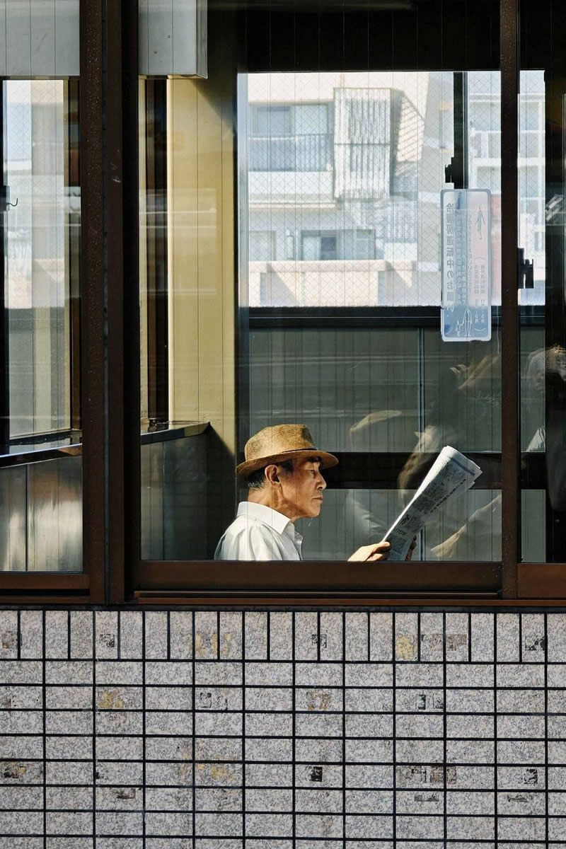 A man wearing a hat sits indoors reading a newspaper, viewed through large window frames. Geometric lines, reflections on the glass, and warm natural light create a layered composition that highlights contemplation, urban life, and visual balance.