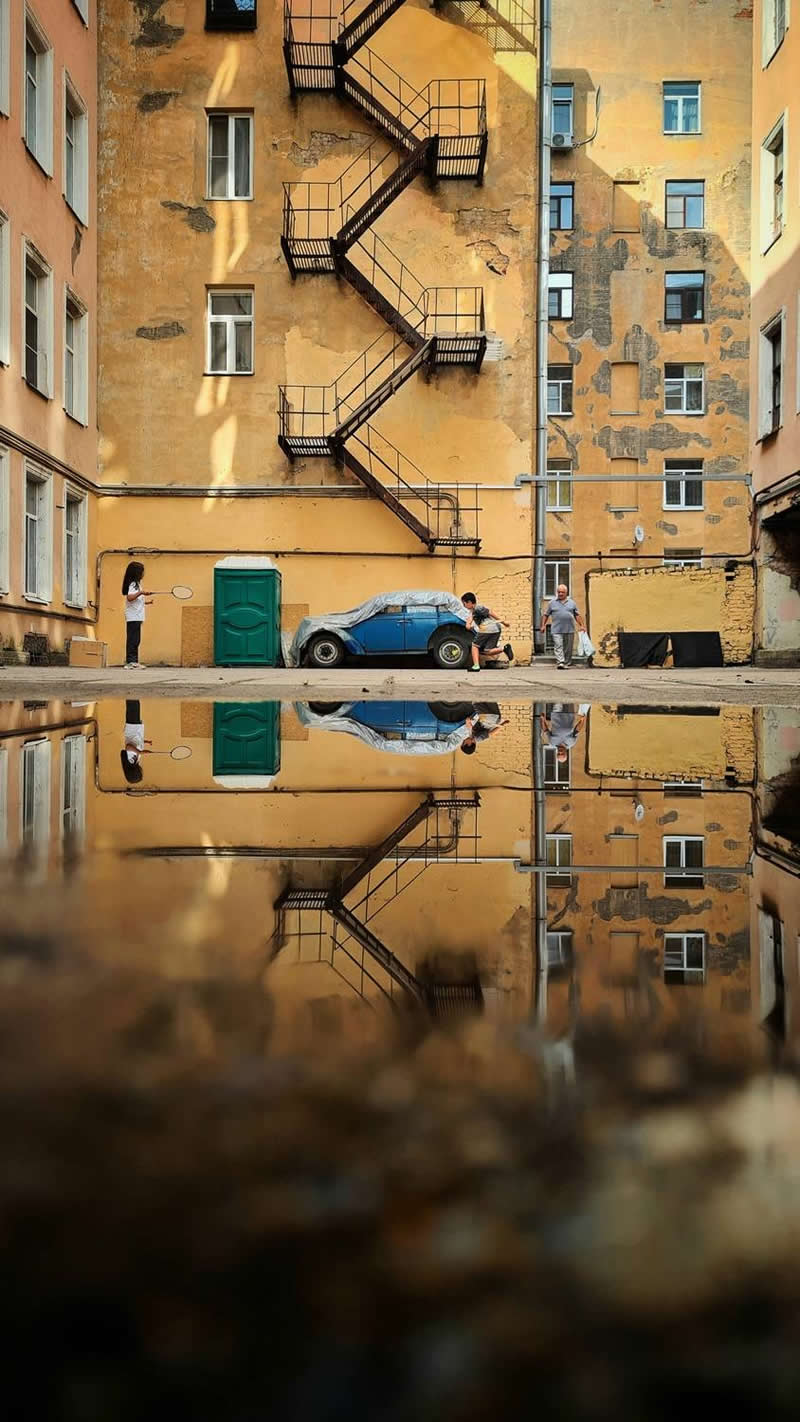 An urban courtyard surrounded by aging apartment buildings features a zigzag fire escape staircase, a parked blue car, and several people. A large puddle in the foreground reflects the entire scene, creating strong symmetry, depth, and a visually captivating composition.