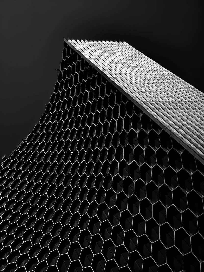 A dramatic low-angle view of a modern high-rise building featuring a repeating honeycomb-patterned facade. The geometric design rises toward a dark sky, emphasizing scale, symmetry, texture, and architectural elegance through strong contrast and striking perspective.