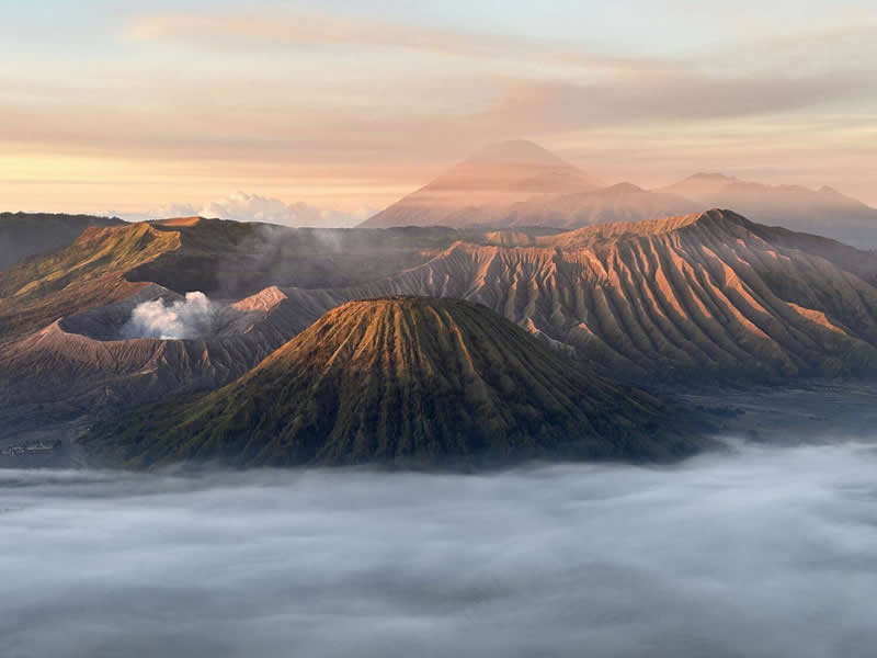 A panoramic view of volcanic mountains rising above a blanket of clouds during sunrise. Warm golden light highlights the ridged slopes, while distant peaks fade into the horizon. The layered landscape creates a dramatic sense of scale, depth, and natural beauty.