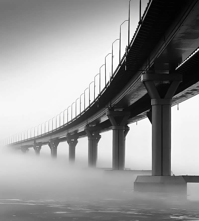 A long bridge curves gracefully across a fog-covered body of water in a black-and-white scene. Repeating support columns and railings create strong leading lines, while dense mist obscures the distance, adding atmosphere, depth, and a sense of mystery.