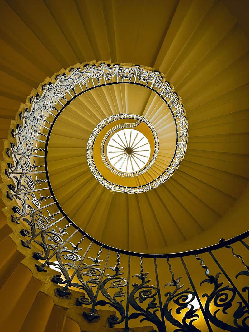 An upward view of a grand spiral staircase with ornate wrought-iron railings and warm golden tones. The steps curve inward toward a bright circular center, creating striking symmetry, depth, and a captivating geometric pattern that emphasizes architectural beauty.