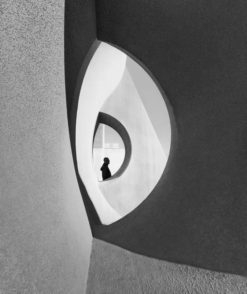 A silhouetted person stands within a circular opening framed by larger curved architectural shapes in a black-and-white scene. Smooth textures, flowing lines, and strong contrast create a minimalist composition emphasizing depth, symmetry, and the relationship between space and scale.