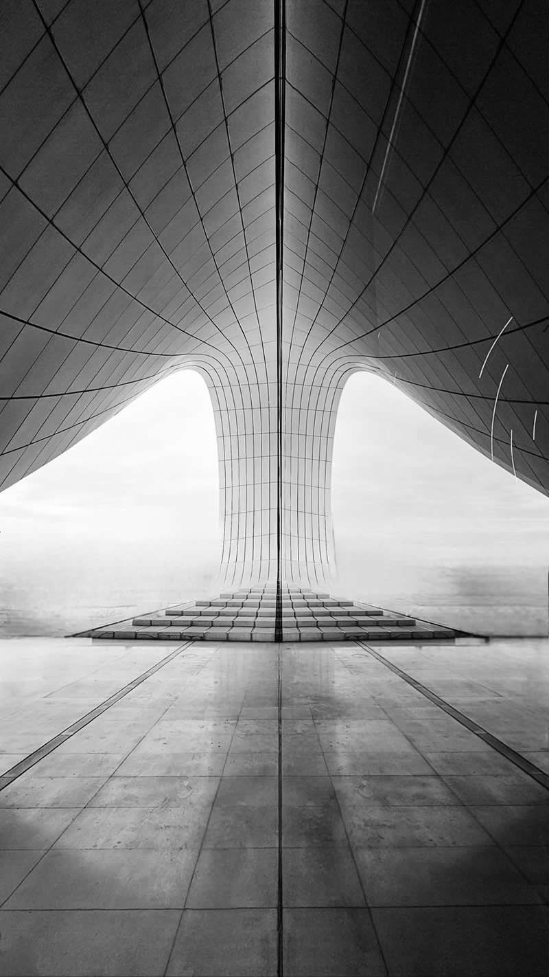 A black-and-white architectural scene viewed from a perfectly centered perspective. Smooth curved surfaces and geometric lines converge toward a central structure, creating striking symmetry, depth, and scale. The minimalist composition emphasizes form, balance, and the beauty of contemporary architecture.