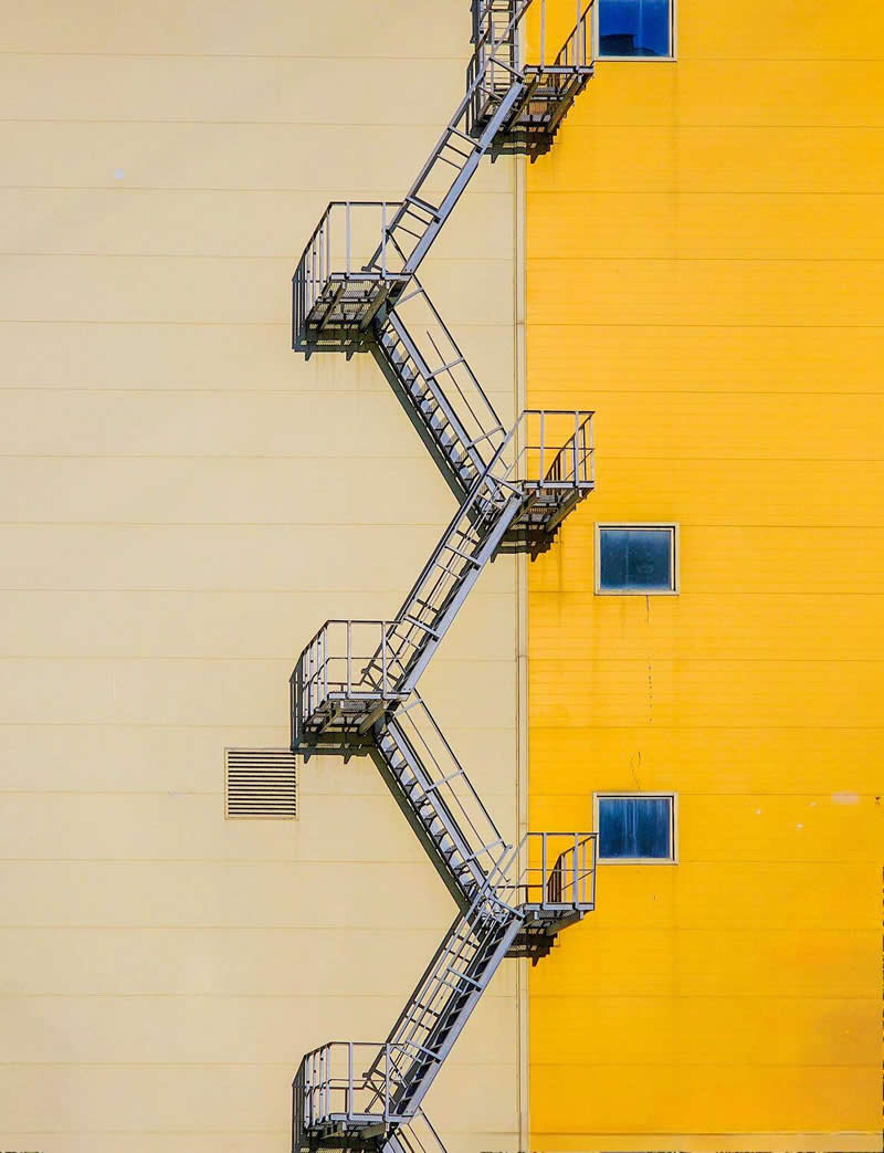 A metal fire escape staircase zigzags vertically across the junction of two building walls, one cream-colored and the other bright yellow. Small windows and clean geometric lines create a minimalist architectural composition emphasizing symmetry, contrast, pattern, and visual balance.