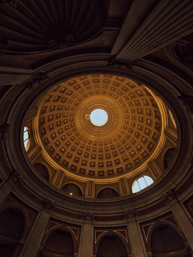 An upward view of a grand domed ceiling inside a historic building, featuring intricate geometric patterns, decorative arches, and classical columns. Warm golden light highlights the architectural details, while a circular skylight at the center creates symmetry, depth, and visual balance.