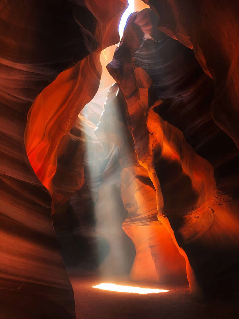 A narrow sandstone canyon glows with rich orange and red hues as a powerful beam of sunlight streams through an opening above. The illuminated dust and flowing rock formations create a dramatic, ethereal scene filled with depth, texture, and natural beauty.