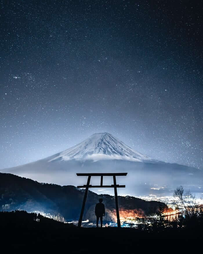 A lone person stands beneath a traditional torii gate, gazing toward snow-capped Mount Fuji beneath a star-filled night sky with distant city lights glowing below.