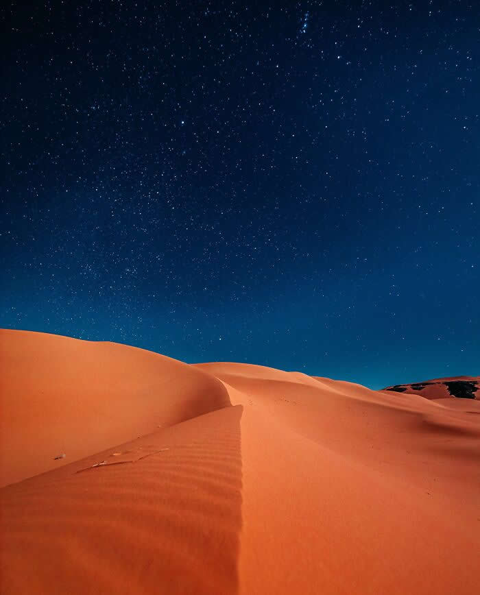 Smooth orange sand dunes illuminated beneath a clear, star-filled night sky, with elegant ridges and sweeping curves creating a minimalist desert landscape.