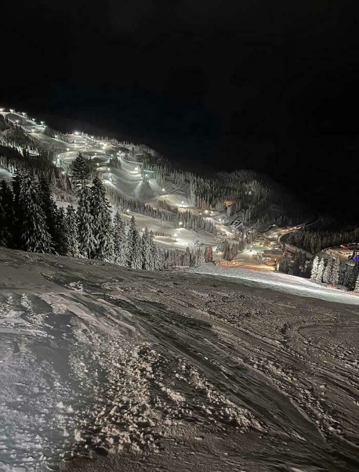 An illuminated ski resort at night with glowing snow-covered slopes, evergreen trees, and textured snowy terrain in the foreground beneath a dark winter sky.