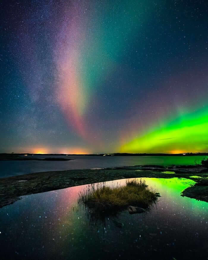 Colorful northern lights illuminate a starry night sky above a calm lake, with vivid green and pink auroras reflected in still water surrounding a small grassy island.