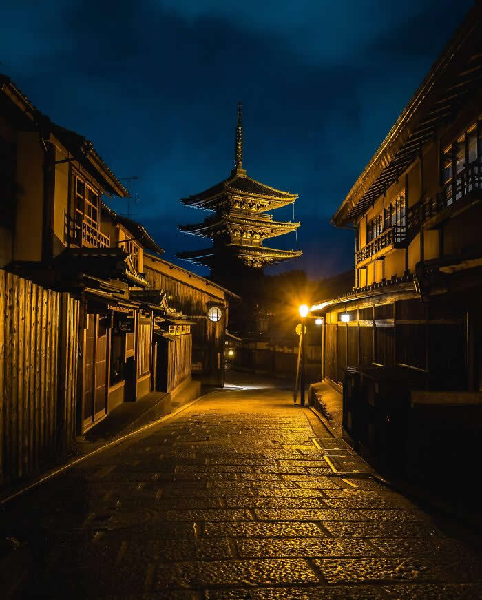 A quiet historic Japanese street at night, illuminated by warm lanterns, leading toward a beautifully lit five-story pagoda beneath a deep blue evening sky.