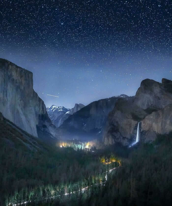 A star-filled night sky stretches above a dramatic mountain valley with towering granite cliffs, a glowing waterfall, winding river, and distant lights illuminating the forest below.