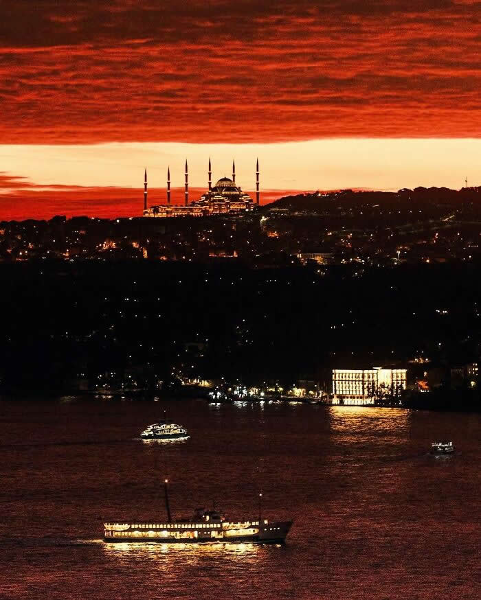 A brilliantly lit mosque stands beneath a dramatic crimson sunset, overlooking a waterfront where illuminated ferries glide across calm water and city lights sparkle in the distance.