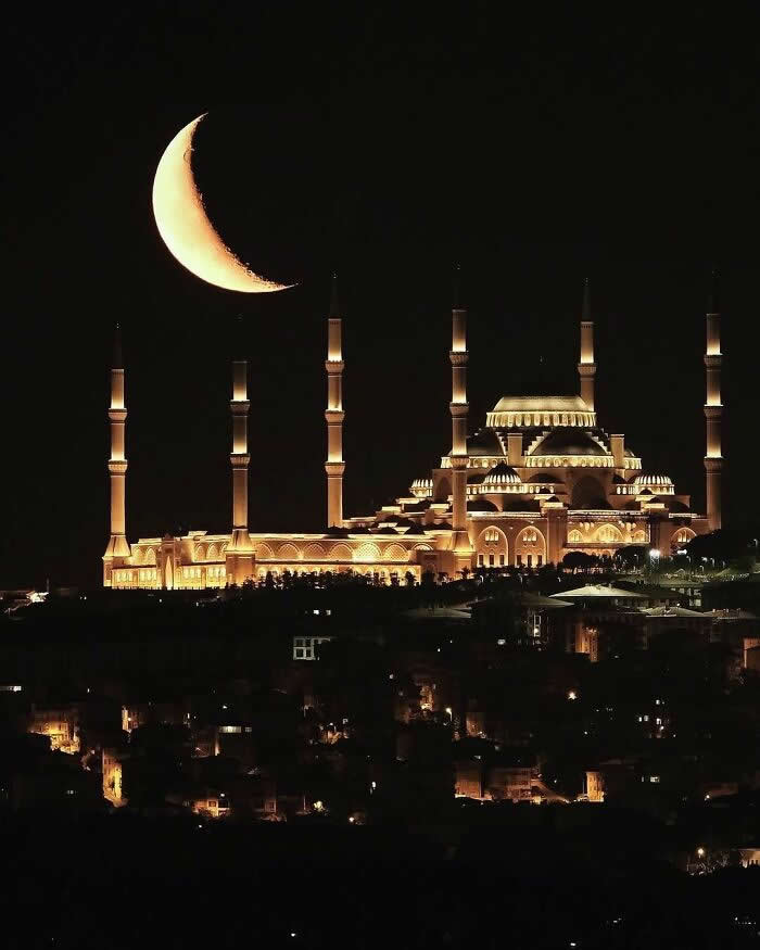 A glowing crescent moon hangs above a brilliantly illuminated mosque with towering minarets, rising over a dark city skyline under a clear night sky.