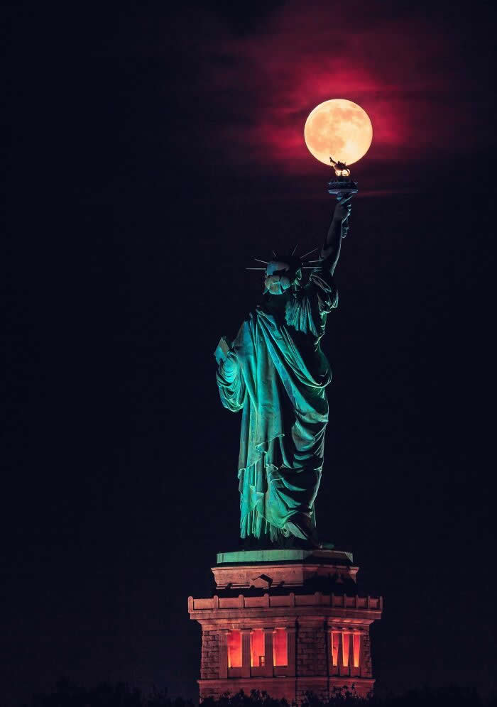 The Statue of Liberty at night appears to hold a glowing full moon atop its torch, creating a striking visual alignment against a dark sky with crimson-tinted clouds.
