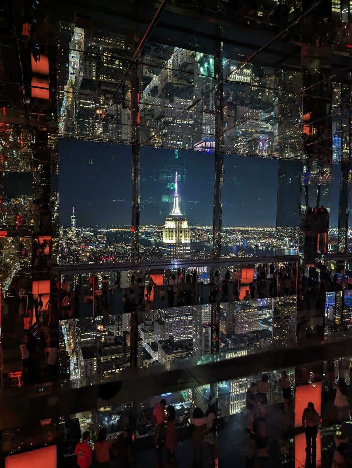 A nighttime city skyline reflected across mirrored walls inside an observation deck, with a brightly illuminated skyscraper centered among endless reflections and visitors silhouetted below.