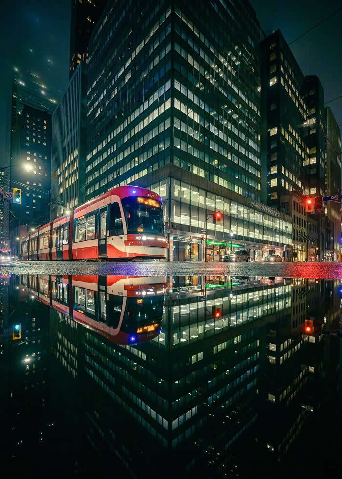 A red streetcar travels through a modern downtown intersection at night, reflected perfectly in a rain puddle beneath illuminated office buildings and colorful city lights.