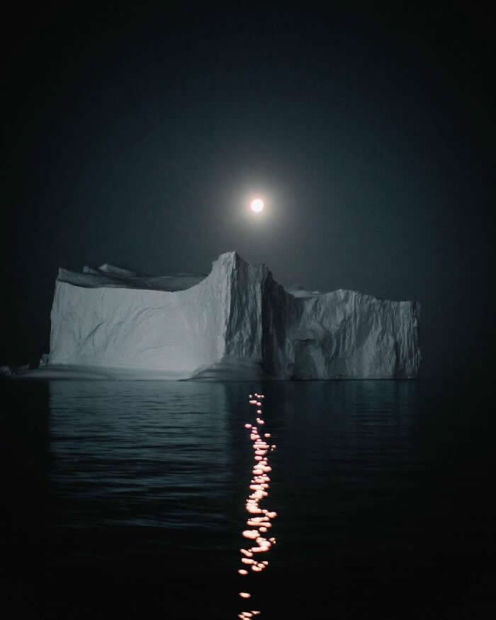 A massive iceberg floats on calm dark water beneath a glowing full moon, with shimmering moonlight reflecting across the surface under a peaceful night sky.