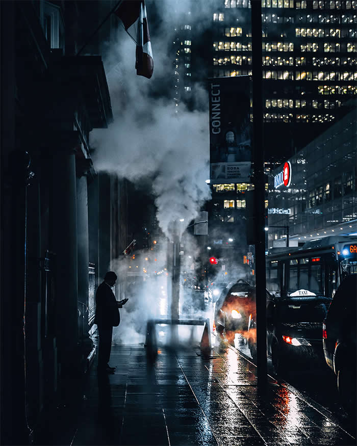A lone person stands on a rain-slicked city sidewalk at night as steam billows from a street vent, surrounded by glowing traffic, illuminated office buildings, and reflections on wet pavement.