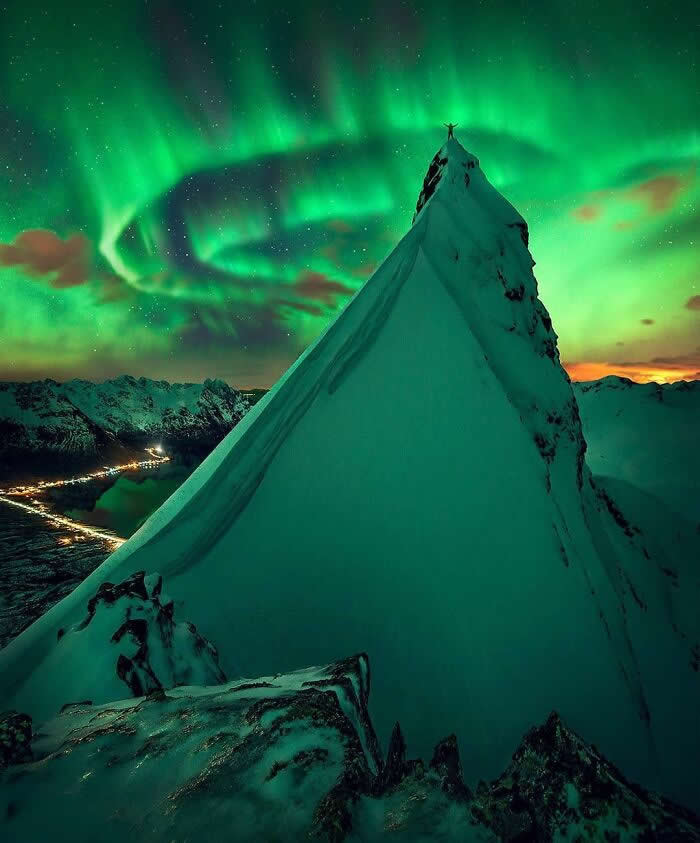 A snow-covered mountain peak rises beneath vibrant green northern lights, with a lone climber standing at the summit overlooking a dramatic winter landscape at night.
