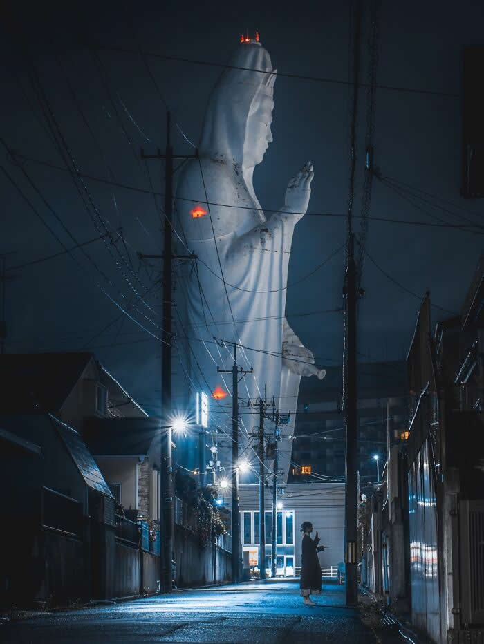 A giant illuminated statue towers above a quiet residential street at night, while a lone person stands beneath glowing streetlights surrounded by utility poles and overhead wires.