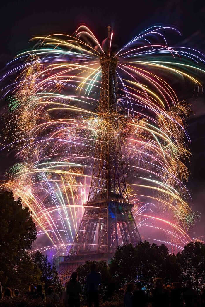 Colorful fireworks explode around the Eiffel Tower at night, filling the sky with vibrant light trails as silhouetted spectators watch the dazzling celebration below.