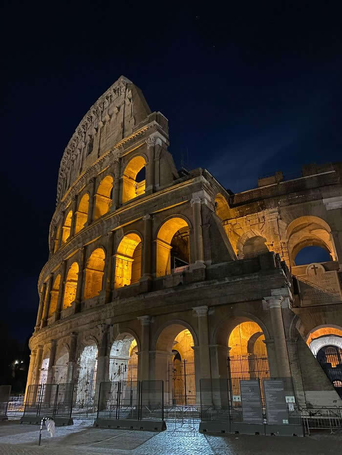 The illuminated Colosseum at night viewed from a low angle, with glowing arches, ancient stone architecture, and a dark blue sky highlighting Rome's iconic historic landmark.