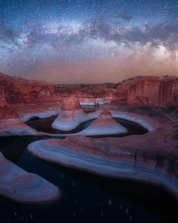 The Milky Way stretches across a star-filled night sky above smooth sandstone rock formations and calm water, creating a peaceful desert landscape with layered canyon walls.