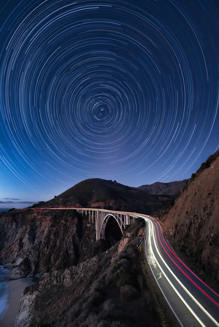 Circular star trails fill the night sky above a dramatic coastal bridge, while long-exposure vehicle light trails curve along a winding cliffside highway overlooking the ocean.