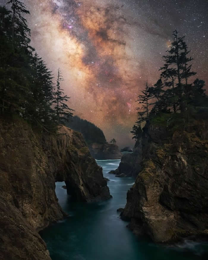 The Milky Way shines above rugged coastal cliffs and a natural sea arch, with calm blue water flowing through the rocky cove beneath a star-filled night sky.