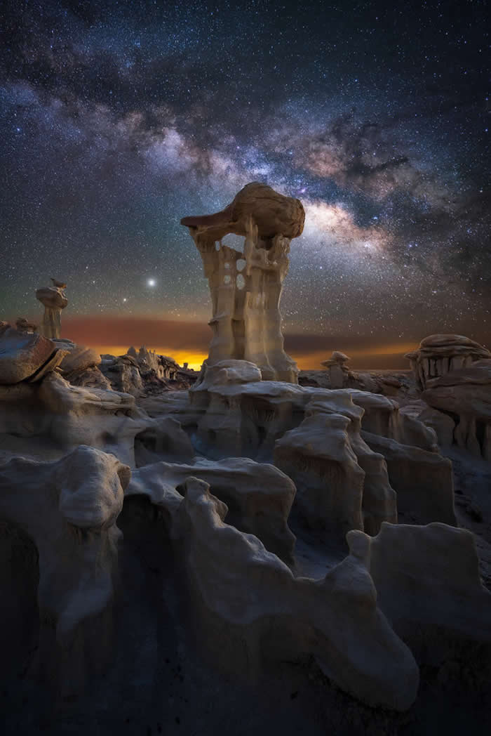 Unique hoodoo rock formations stand beneath the Milky Way on a clear night, with rugged desert terrain illuminated by faint horizon light and thousands of stars overhead.