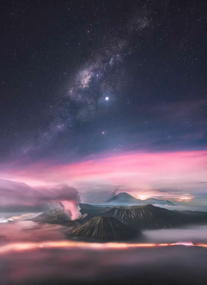 The Milky Way stretches above steaming volcanic mountains surrounded by mist, with glowing pink clouds and layered peaks creating a dramatic nighttime volcanic landscape.