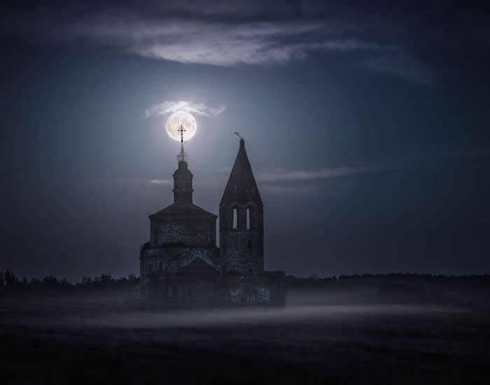 An old abandoned church stands in a misty field at night, with a glowing full moon perfectly aligned above its cross beneath a dark, cloud-streaked sky.