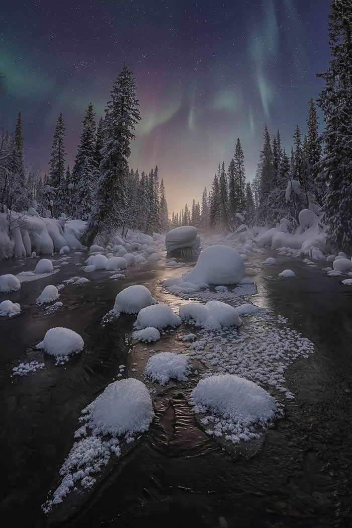 A peaceful snow-covered river winds through a forest beneath the northern lights, with frosted rocks, tall pine trees, and a star-filled winter sky creating a serene nighttime landscape.