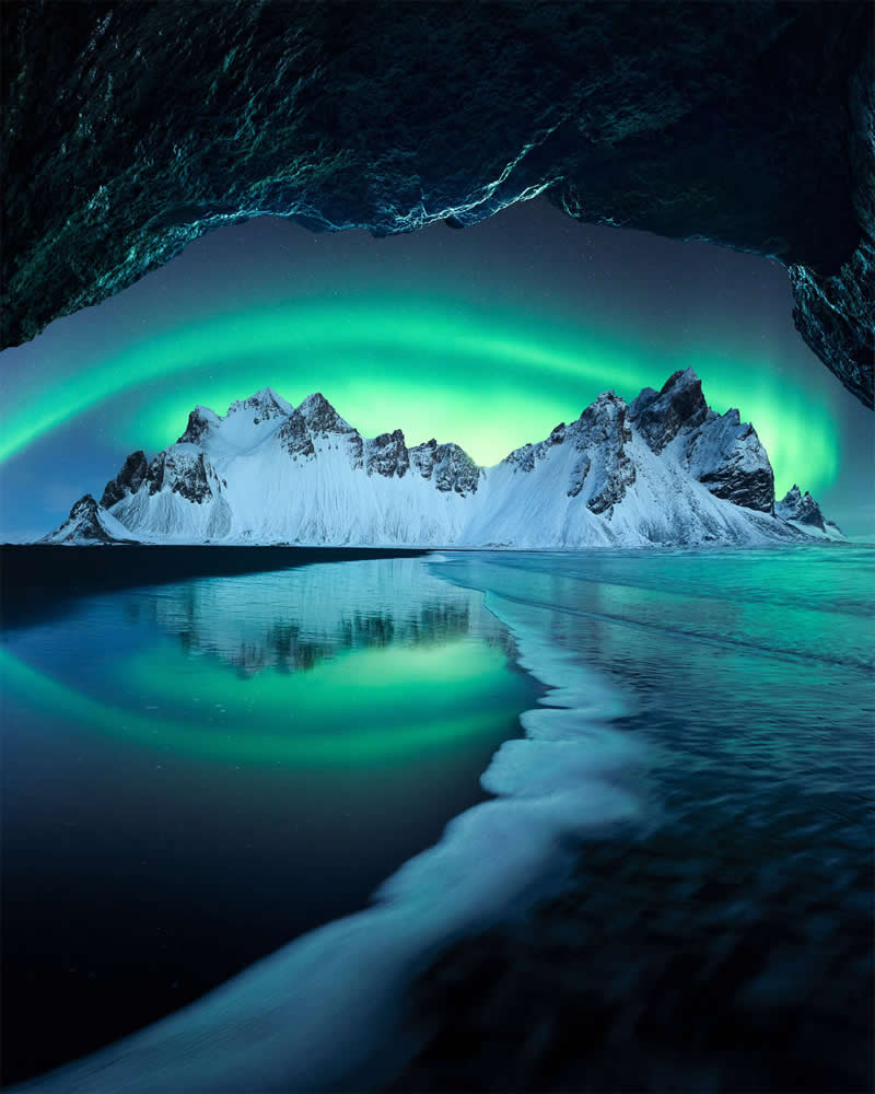 The Northern Lights illuminate snow-covered mountains in Iceland, reflected in calm water and beautifully framed by the opening of a dark sea cave, creating a stunning landscape photograph.