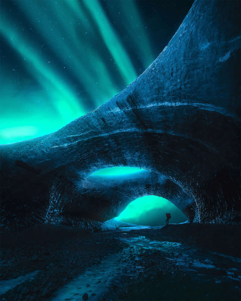 A lone photographer stands beneath towering blue ice arches in Iceland as the green Northern Lights illuminate the night sky, creating a dramatic and beautifully framed Arctic landscape.