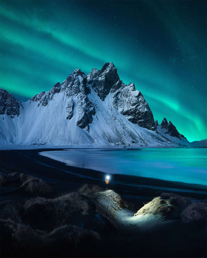 A lone photographer with a headlamp stands before Iceland's snow-covered mountains beneath the glowing Northern Lights, creating a dramatic nighttime landscape with stunning composition and Arctic scenery.