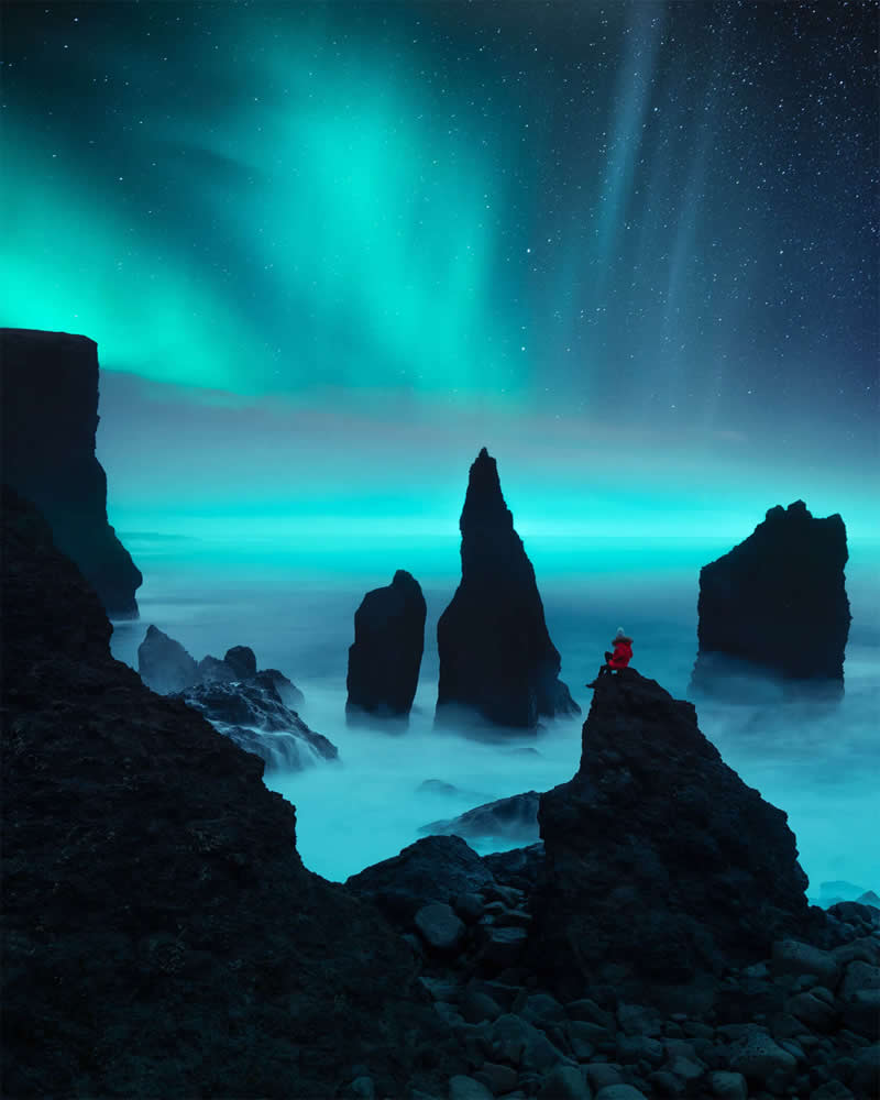 A lone person in a red jacket sits on a rocky sea stack beneath the glowing Northern Lights, surrounded by Iceland's dramatic coastal rock formations and misty ocean at night.