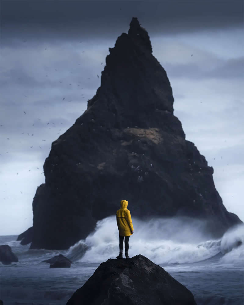 A lone person in a yellow rain jacket stands on a rock facing a towering black sea stack in Iceland as powerful ocean waves crash beneath a dramatic overcast sky.