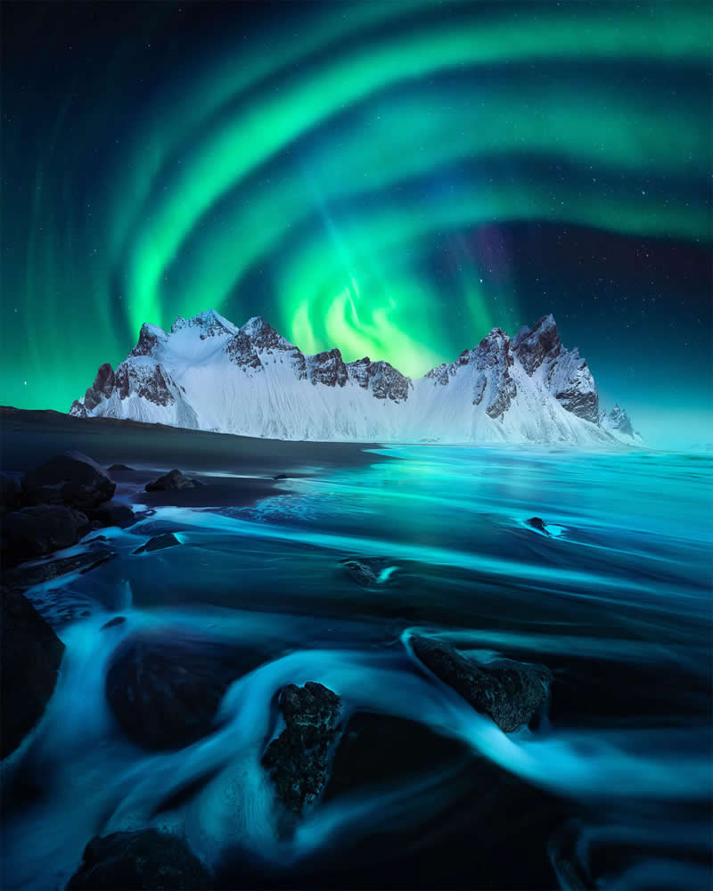 The Northern Lights swirl above Iceland's snow-covered mountains as smooth waves wash over a black volcanic beach, creating a dramatic nighttime landscape with vibrant green auroras and striking reflections.