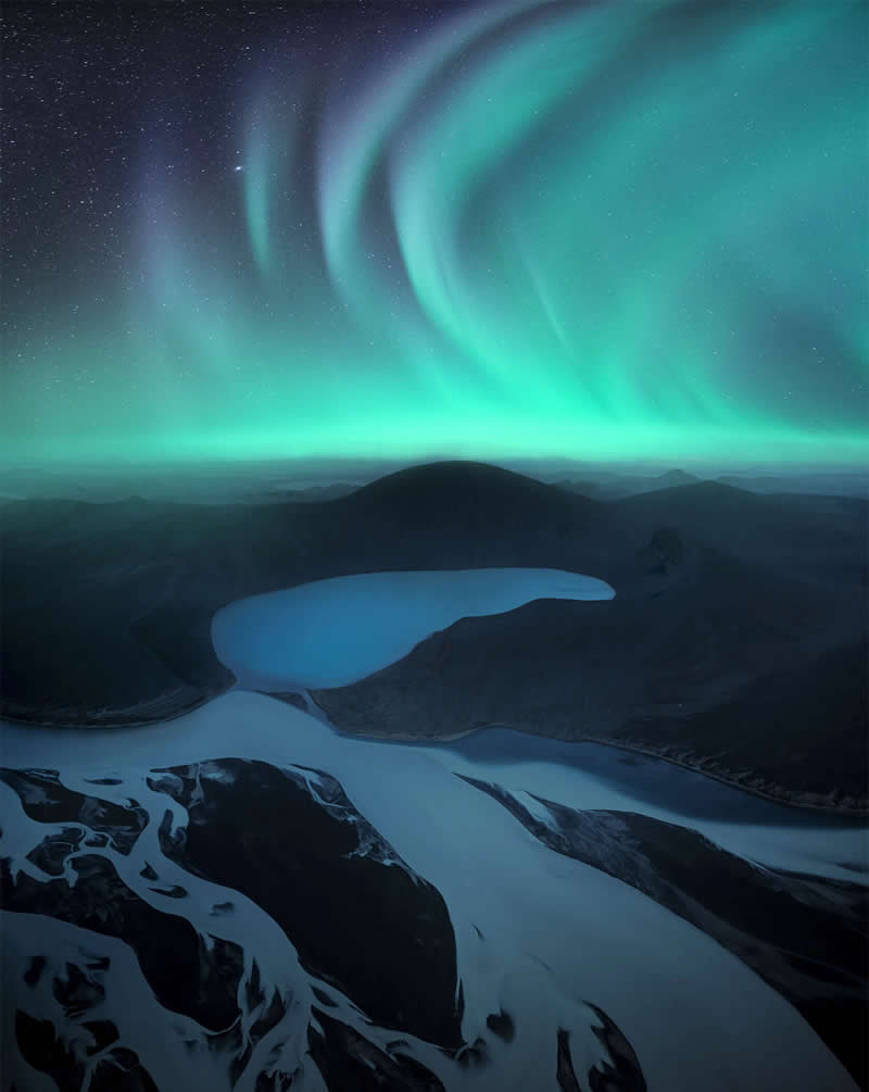 The Northern Lights glow above Iceland's volcanic mountains and tranquil lakes, viewed from a high snowy ridge under a star-filled sky, creating a serene and dramatic Arctic landscape.
