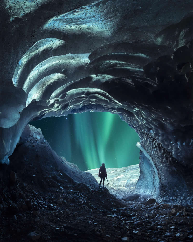 A lone explorer stands inside a blue ice cave in Iceland, framed by sculpted glacier walls while the glowing Northern Lights illuminate the cave entrance beneath a star-filled Arctic sky.