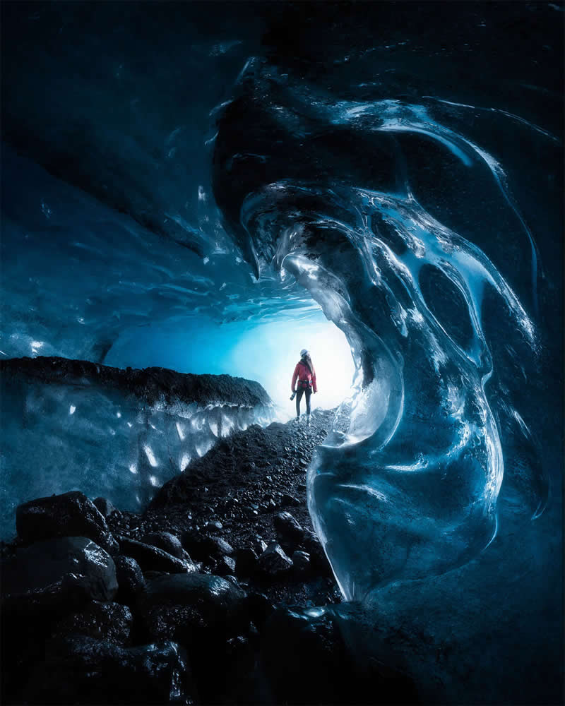 A lone explorer stands inside a crystal-blue ice cave in Iceland, framed by sculpted glacier formations and glowing light at the cave entrance, creating a dramatic and artistic Arctic landscape.