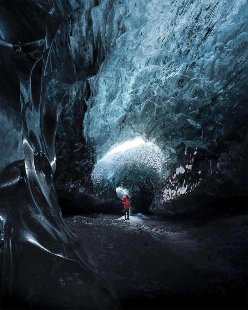 A lone explorer stands inside a massive blue glacier cave in Iceland, surrounded by towering ice walls and illuminated by soft natural light streaming through the cave entrance.